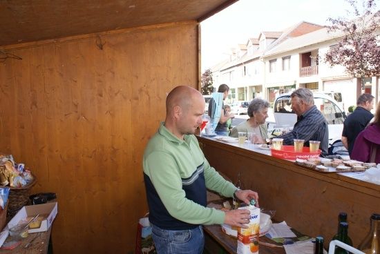 Ein Mann in einem grünen Shirt steht an einer Theke und bereitet Essen oder Getränke in einem hölzernen Raum zu, mit anderen Personen im Hintergrund.