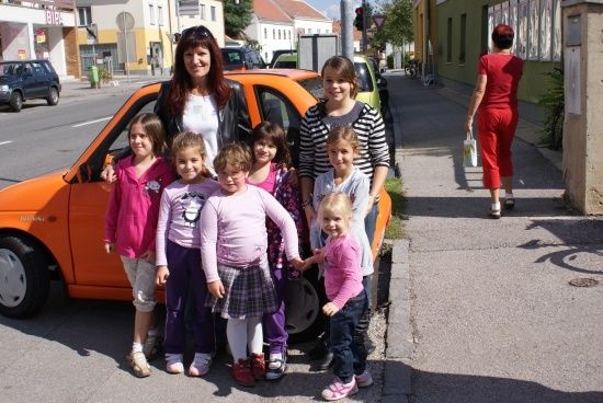 Eine Frau und mehrere Kinder stehen neben einem orangefarbenen Auto auf einer Straße.