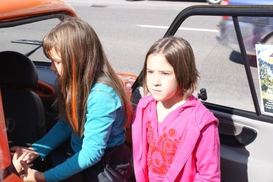 Zwei junge Mädchen, eines in einem blauen Shirt und das andere in einer rosa Jacke, stehen neben einem orangefarbenen Auto. Das Mädchen in Blau sitzt im Auto, während das andere Mädchen draußen steht.