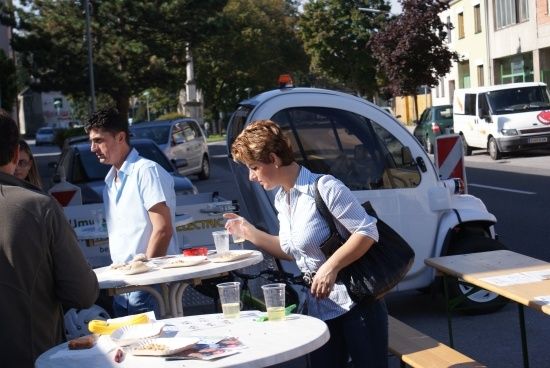 Eine Frau steht an einem Tisch mit Essen und Getränken, während ein Mann sie ansieht. Hinter ihnen ist ein weißer Elektrowagen am Straßenrand geparkt.