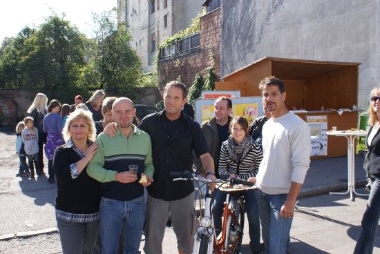 Eine Gruppe von Menschen steht auf der Straße, einige mit Fahrrädern, und andere gehen hinter ihnen. Im Hintergrund befinden sich Gebäude und Bäume.