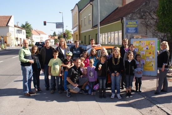 Eine Gruppe von Menschen, einschließlich Erwachsener und Kindern, posiert für ein Foto an einer Straßenecke. Dahinter steht ein Auto und ein Schild. Gebäude und Bäume sind im Hintergrund.