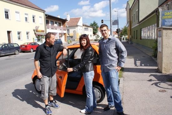Drei Personen stehen neben einem orangefarbenen Auto auf einer Stadtstraße, mit Gebäuden und geparkten Autos im Hintergrund.