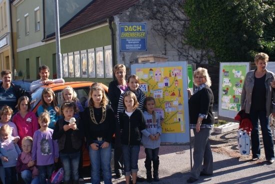 Eine Gruppe von Kindern und Erwachsenen steht draußen und lächelt für ein Foto. Sie stehen vor einem Gebäude mit einem Schild, das Dach Ehrenberger liest. Die Kinder halten Eiswaffeln.