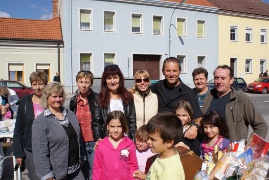 Eine Gruppe von Menschen, einschließlich Erwachsener und Kindern, steht zusammen vor einem Gebäude mit mehreren Fenstern und einem roten Dach. Sie lächeln und posieren für ein Foto.