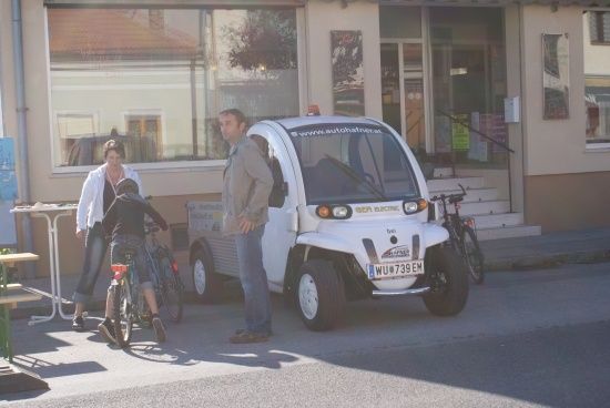 Ein Mann und zwei Kinder stehen vor einem kleinen weißen Elektroauto mit einem Nummernschild. Die Kinder fahren Fahrräder, und der Mann trägt eine Jacke. Dahinter befindet sich ein Gebäude mit Glasfenstern und einer Treppe.