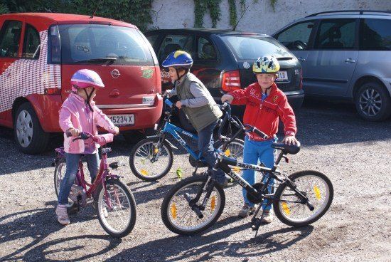 Drei Kinder stehen mit ihren Fahrrädern auf einem Parkplatz. Das Mädchen links trägt eine rosa Jacke, während der Junge rechts eine rote Jacke trägt. Hinter ihnen sind drei Autos geparkt.