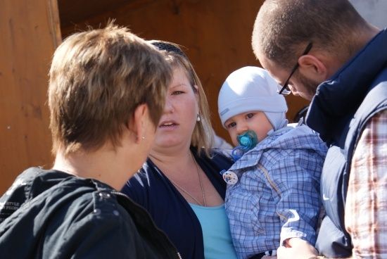 Eine Familie mit einem Baby, das einen Schnuller im Mund hat, das Baby trägt eine blaue Jacke und eine weiße Mütze.