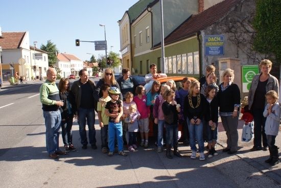 Eine Gruppe von Erwachsenen und Kindern steht an einer Straßenecke, einige halten Eiscreme, mit Gebäuden und einer Ampel im Hintergrund.