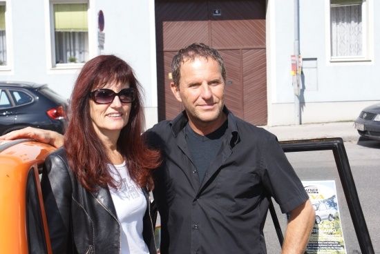 Ein Mann und eine Frau stehen gemeinsam vor einem Gebäude mit geparkten Autos und einer hölzernen Tür.