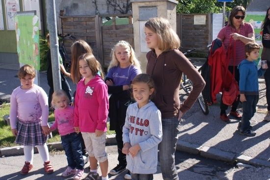 Eine Gruppe von Kindern und Erwachsenen steht auf einem Bürgersteig, einige halten sich an den Händen. Eine Frau in einem braunen Shirt hält ein Fahrrad.