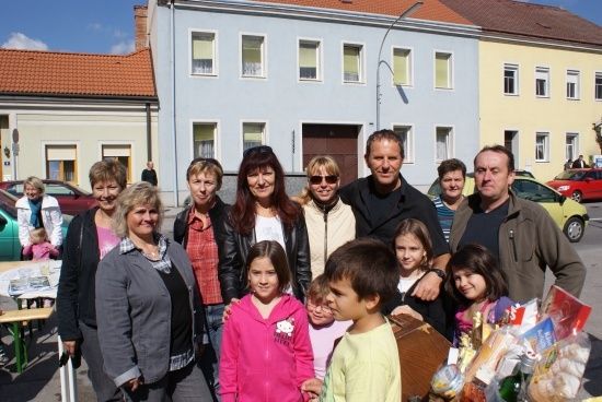 Eine Gruppe von Erwachsenen und Kindern steht vor einem Gebäude mit mehreren Fenstern und geparkten Autos im Hintergrund. Einige lächeln und posieren für ein Foto.