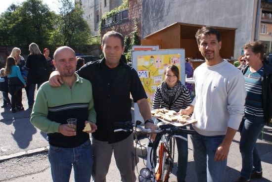 Drei Männer und eine Frau stehen draußen, zwei der Männer halten einen Teller mit Essen und ein Getränk, während der andere Mann ein Fahrrad hält. Hinter ihnen gehen mehrere Leute auf der Straße.