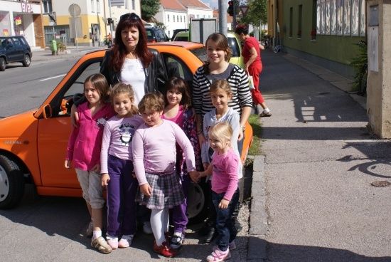 Eine Frau und mehrere junge Mädchen stehen neben einem orangefarbenen Auto auf der Straße.