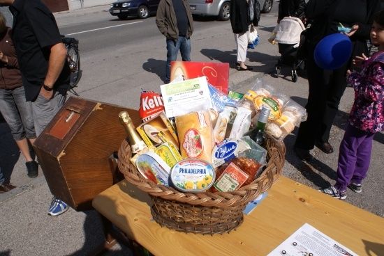 Ein Korb mit verschiedenen Lebensmitteln, einschließlich Brot, Käse und einer Flasche Wein, ist auf einem Holztisch im Freien ausgestellt. Im Hintergrund gehen Menschen vorbei.