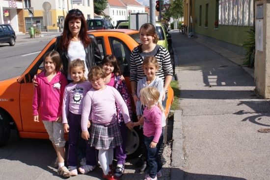 Eine Gruppe von Kindern und zwei Frauen stehen neben einem orangefarbenen Auto auf der Straße. Die Kinder lächeln und scheinen für ein Foto zu posieren.