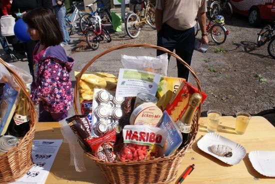 Ein Korb mit verschiedenen Lebensmitteln und Getränken, darunter eine Flasche Wein, Haribo-Süßigkeiten und andere Snacks, steht auf einem Holztisch. Ein Mädchen in einer pinken Jacke steht in der Nähe, und im Hintergrund sind Fahrräder geparkt.