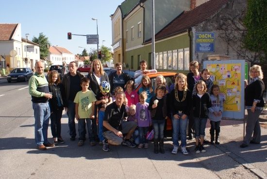 Eine Gruppe von Menschen, einschließlich Erwachsener und Kinder, steht am Straßenrand vor einem Gebäude. Einige tragen Helme und Turnschuhe.