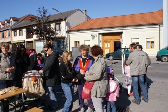Eine Gruppe von Menschen, darunter Frauen und Kinder, steht in einer Reihe auf einer Straße vor einem Gebäude. Einige halten Gegenstände, und ein Kinderwagen ist in der Nähe.