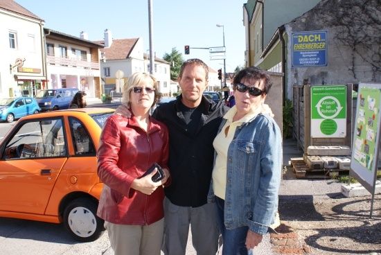 Drei Personen stehen zusammen an einer Straßenecke vor einem orangefarbenen Auto. Eine Frau in einer roten Jacke und ein Mann in einer schwarzen Jacke stehen zwischen zwei Frauen mit Sonnenbrillen. Hinter ihnen befindet sich ein Gebäude mit einem Schild, das Dach steht.