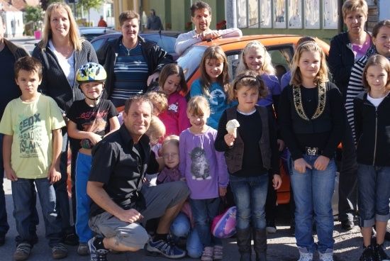 Eine Gruppe von Menschen, einschließlich Erwachsener und Kindern, posiert für ein Foto vor einem Auto. Einige von ihnen tragen Helme und Turnschuhe.