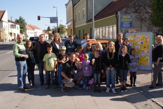 Eine Gruppe von Menschen, einschließlich Erwachsener und Kindern, posiert für ein Foto an einer Straßenecke. Einige tragen Helme und Turnschuhe. Ein Auto ist in der Nähe geparkt.