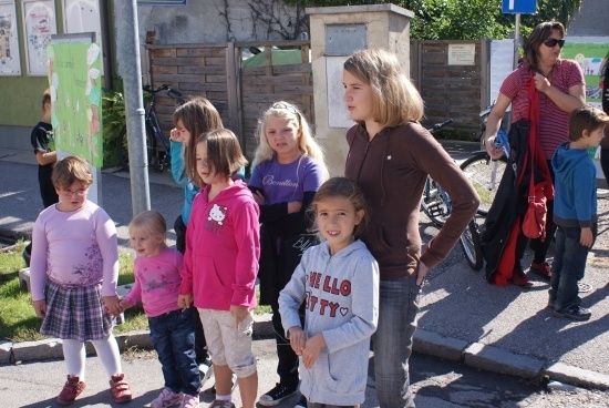 Eine Gruppe von Kindern und Erwachsenen steht auf einem Bürgersteig, mit geparkten Fahrrädern und einem grünen Schild im Hintergrund.