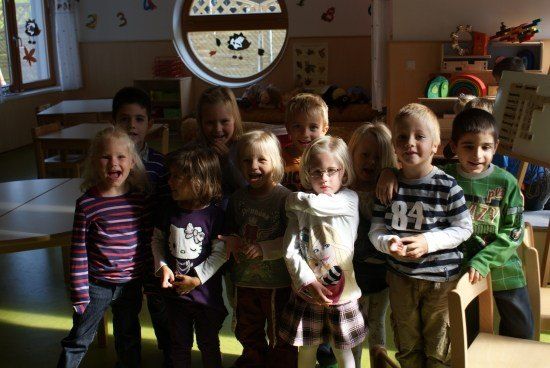 Eine Gruppe junger Kinder, einige mit Brille, steht in einem Klassenzimmer zusammen, mit Tischen und Stühlen im Hintergrund.