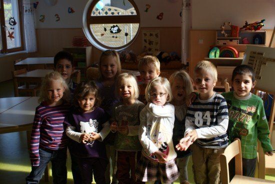 Eine Gruppe von Kindern in einem Klassenzimmer, die lächeln und für ein Foto posieren. Sie stehen in einer Reihe, einige halten Spielzeug. Hinter ihnen sind Tische, Stühle und ein rundes Fenster.