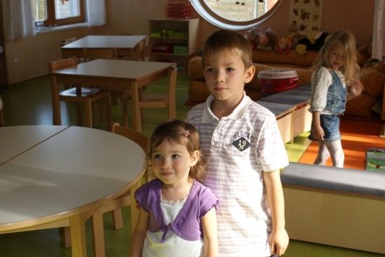 Zwei Kinder, ein Junge und ein Mädchen, stehen in einem Raum mit Möbeln, einer Couch und einem Fenster.