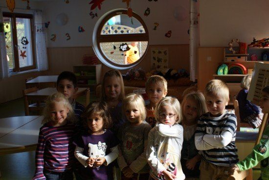 Eine Gruppe junger Kinder, wahrscheinlich in einem Klassenzimmer, posiert für ein Foto. Sie stehen eng beieinander, und einige lächeln. Der Raum hat weiße Wände, ein kreisförmiges Fenster und verschiedene Dekorationen.