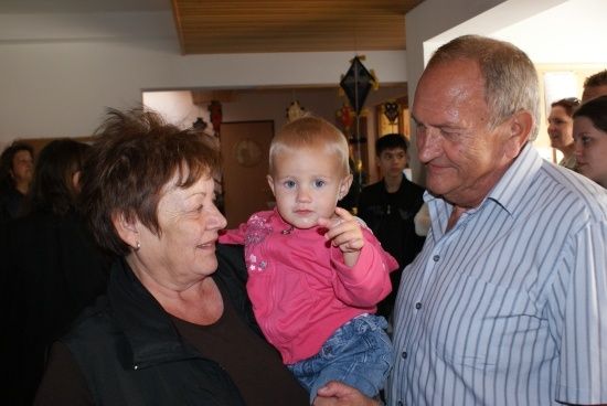 Ein älteres Paar hält ein Kleinkind in einem Raum, mit anderen Personen im Hintergrund.