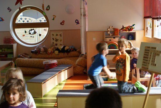 Eine Gruppe von Kindern spielt in einem Spielzimmer mit Spielzeug und Möbeln. Ein Junge springt, während ein anderer Junge auf dem Boden sitzt.