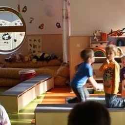 Eine Gruppe von Kindern spielt in einem Spielzimmer mit Spielzeug und Möbeln. Ein Junge springt, während ein anderer Junge auf dem Boden sitzt.