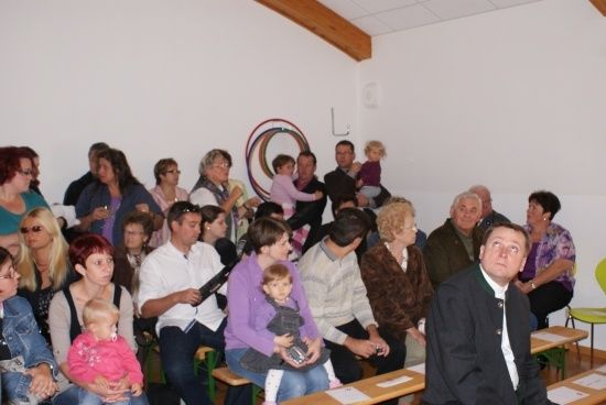 Eine Gruppe von Menschen sitzt in einem Raum, einige halten Babys. Ein Mann in Anzug steht vor ihnen. Es gibt Holztische und Stühle. Ein kreisförmiges Objekt ist an der Wand befestigt.