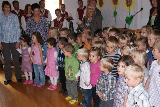 Eine Gruppe von Kindern und Erwachsenen ist drinnen versammelt, wahrscheinlich für eine Schulveranstaltung. Die Kinder stehen in einer Reihe, und einige halten Händchen. Hinter ihnen spricht eine Frau in ein Mikrofon. Der Raum hat Holzböden und eine weiße Wand mit Sonnenblumen.