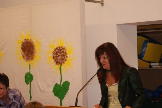Eine Frau steht an einem Podium und spricht in ein Mikrofon mit einem Sonnenblumenhintergrund. Eine weitere Person sitzt im Hintergrund und trägt eine Brille.
