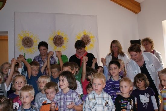 Eine Gruppe von Kindern und Erwachsenen steht in einem Raum mit einem sonnenblumenthematischen Hintergrund. Die Kinder heben ihre Hände, und die Erwachsenen stehen hinter ihnen.