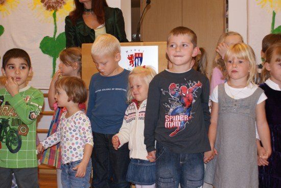 Eine Gruppe von Kindern steht zusammen, einige halten Händchen, und eine Frau steht hinter ihnen. Die Kinder tragen verschiedenfarbige Hemden und Hosen. Der Junge in der Mitte trägt ein Spider-Man-Shirt. Hinter ihnen steht ein Podium mit einem Mikrofon und einem Banner mit Blumendesign.