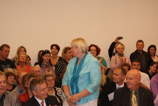 Eine Gruppe von Menschen ist in einem Raum versammelt, einige klatschen, und eine Frau in einem blauen Oberteil steht in der Mitte.