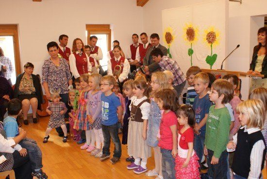 Eine Gruppe von Kindern und Erwachsenen steht in einem Raum mit Holzböden, einige halten Musikinstrumente. Eine Frau steht an einem Podium mit einem Mikrofon, und Sonnenblumen sind an der Wand hinter ihr gemalt.