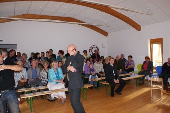 Eine Gruppe von Menschen ist in einem Raum mit Holzböden und Bänken versammelt. Ein Mann in einem Anzug steht vor ihnen und hält möglicherweise eine Rede.