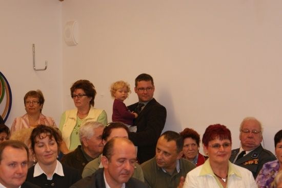 Eine Gruppe von Menschen ist in einem Raum versammelt, einige stehen und einige sitzen. Ein Mann hält ein Kind in den Armen.