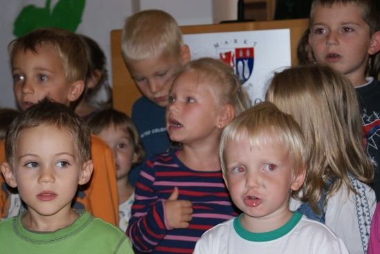 Eine Gruppe von Kindern, einige mit roten Augen, sind in einem Raum versammelt, möglicherweise für eine Schulveranstaltung. Das Mädchen in der Mitte gibt einen Daumen hoch.