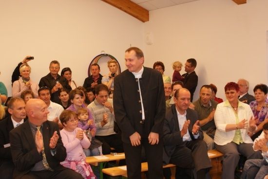 Ein Mann in einem schwarzen Anzug steht vor einer applaudierenden Menschenmenge. Das Publikum sitzt auf Bänken, einige Leute stehen und halten Kameras.
