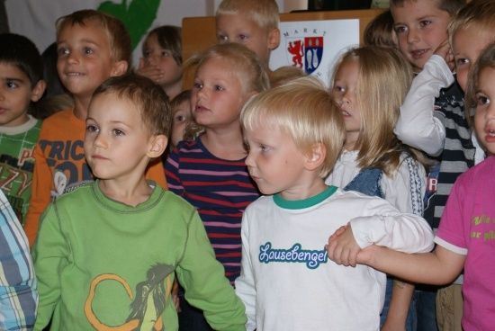 Eine Gruppe junger Kinder, einige in bunten Shirts, stehen in einem Raum zusammen. Der Junge vorne trägt ein grünes Shirt mit einem Cartoon-Charakter.