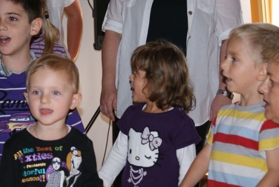 Eine Gruppe von Kindern, darunter ein Junge in einem schwarzen T-Shirt und ein Mädchen in einem lila Hello Kitty-Shirt, stehen zusammen in einem Raum.