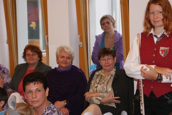 Eine Gruppe von Frauen und einem jungen Mädchen versammelt sich in einem Raum, einige tragen Brillen und lächeln. Ein Mädchen in einer roten Weste hält eine Flöte.