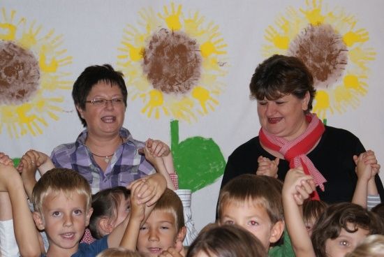 Zwei Frauen und mehrere Kinder stehen vor einer Wand mit handgemalten Sonnenblumen. Die Kinder heben ihre Hände.