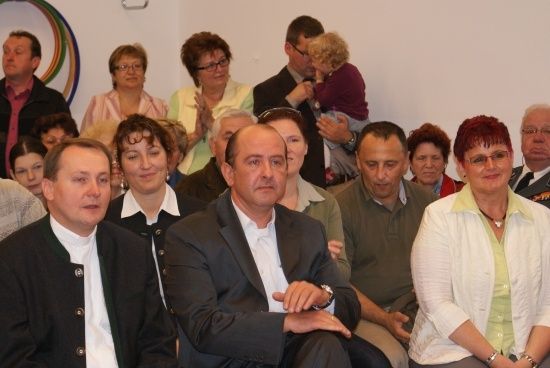 Eine Gruppe von Menschen, einschließlich Erwachsener und Kinder, versammelt sich in einem Raum. Einige sitzen, andere stehen. Ein Mann im Anzug schaut nach vorne.